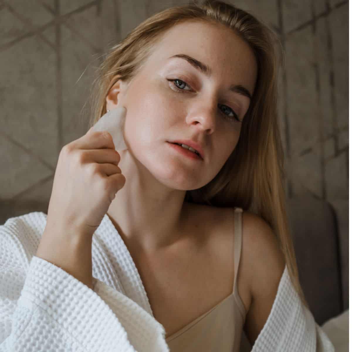 Blonde Woman in White Robe Using Gua Sha On Face